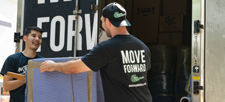 Two professional movers unloading a sofa protected with a moving blanket and tape from a truck showing the benefits of hiring a green moving company in NYC