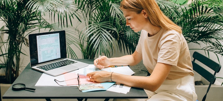 A woman sitting at the table in front of her laptop looking at a pile of documents while preparing for office relocation 