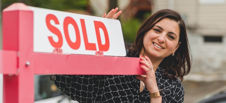 A woman standing next to a sold sign representing moving when you work from home