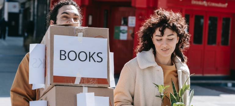 a couple carrying boxes, (one labelled "books") when moving a home library to Park Slope